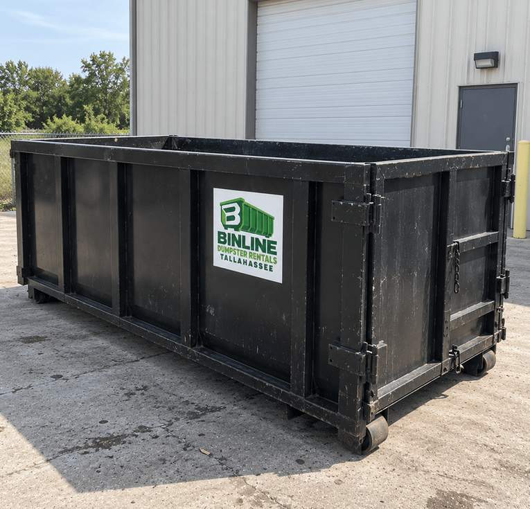 Black open-top roll-off dumpster with the Binline Dumpster Rentals Tallahassee logo, sitting in front of a warehouse door