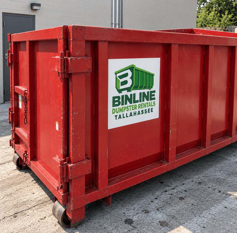 Red steel roll-off dumpster with a hinged rear door and the Binline Dumpster Rentals Tallahassee logo on the side, parked on a concrete yard